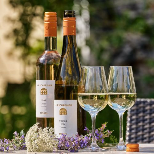 Zwei Flaschen HP Schreiner Wein und zwei gefuellte Weinglaeser mit Blumen auf einem Tisch draussen