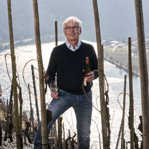 Mann mit Weinflasche und Glas steht im Weinberg vor einem Fluss und Bergen